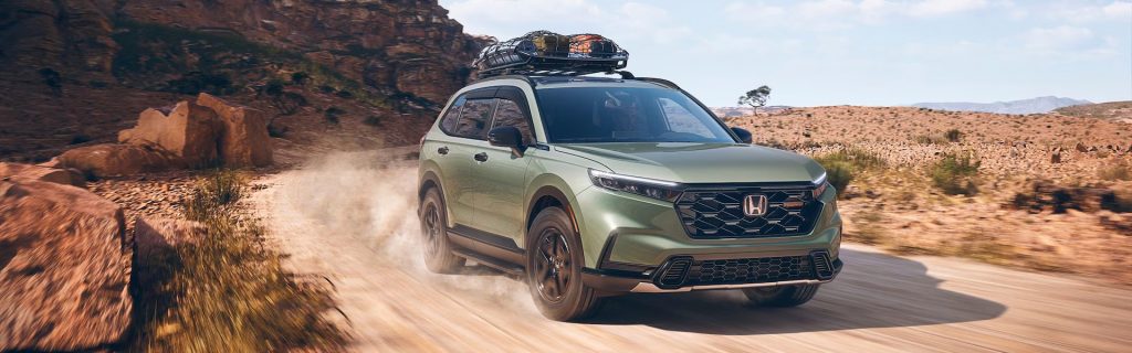 A sage-green Honda SUV kicks up dust while driving along a rugged, desert dirt road with a loaded roof cargo basket.